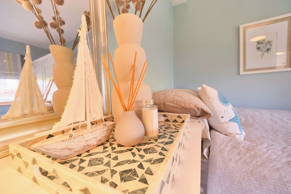 A room with a bed, a decorative mirror, a vase with reed sticks, and a small bottle.
