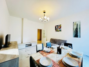 Een eetgedeelte met een tafel voor vier personen, een bank, een televisie en kunstwerken aan de muur.