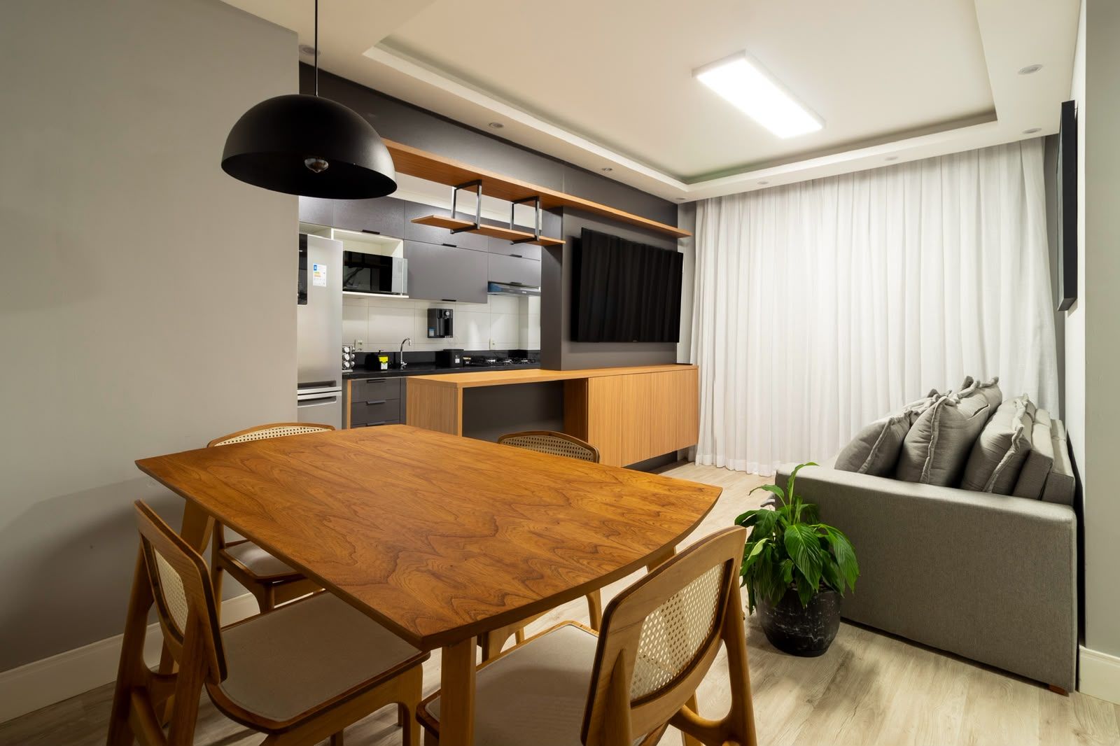 A modern kitchen with built-in appliances, a dining area with a wooden table and chairs, and a grey sofa in a living area.