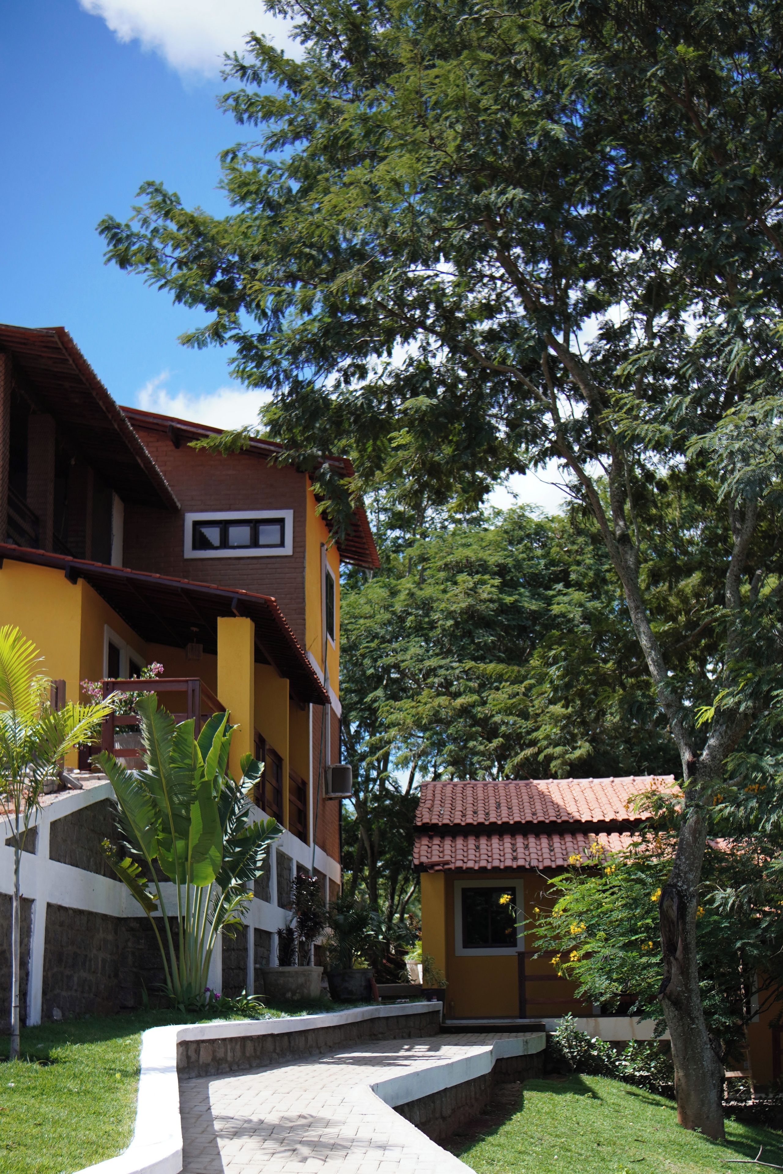 A two-story building with a yellow facade and a brown roof, surrounded by lush greenery and a clear blue sky.