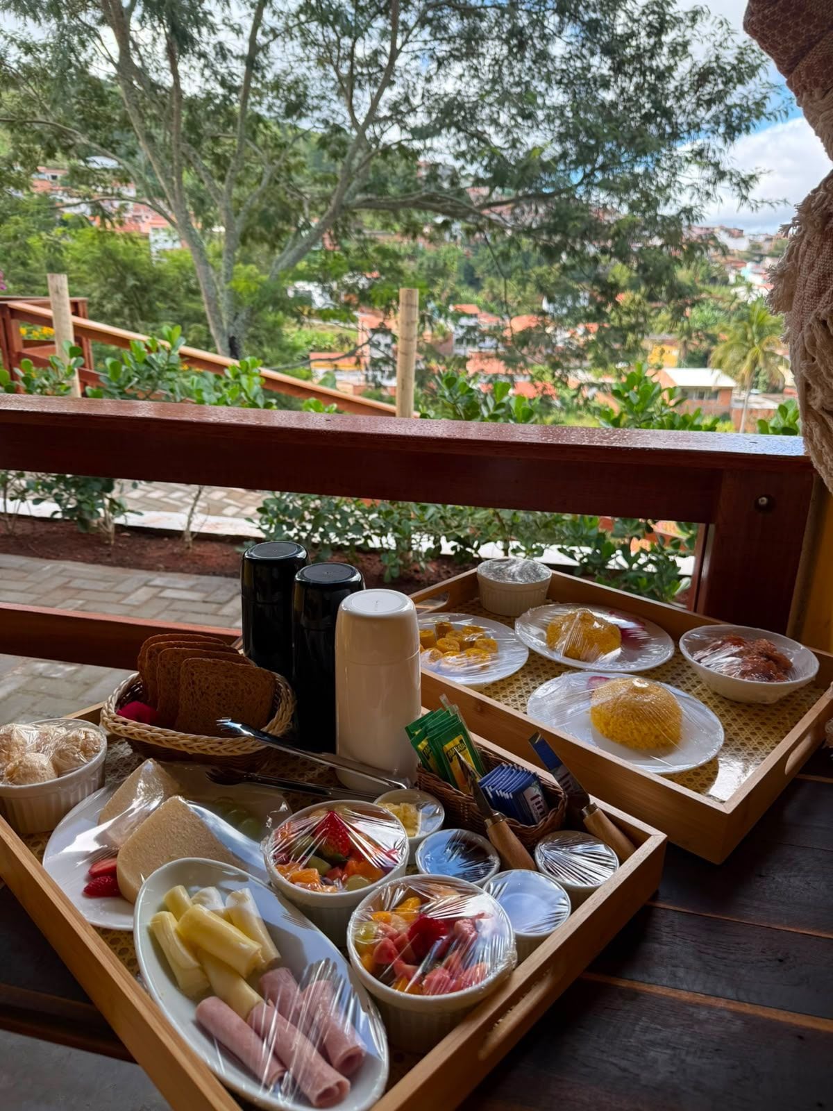Un bufé de desayuno con diversos alimentos y condimentos sobre bandejas de madera, situado en un balcón con vistas a una zona residencial.