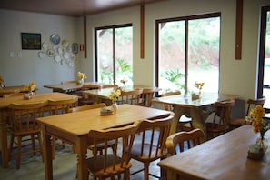 Un comedor con mesas y sillas de madera, una pared decorada con platos, y ventanas amplias con vista a follaje.