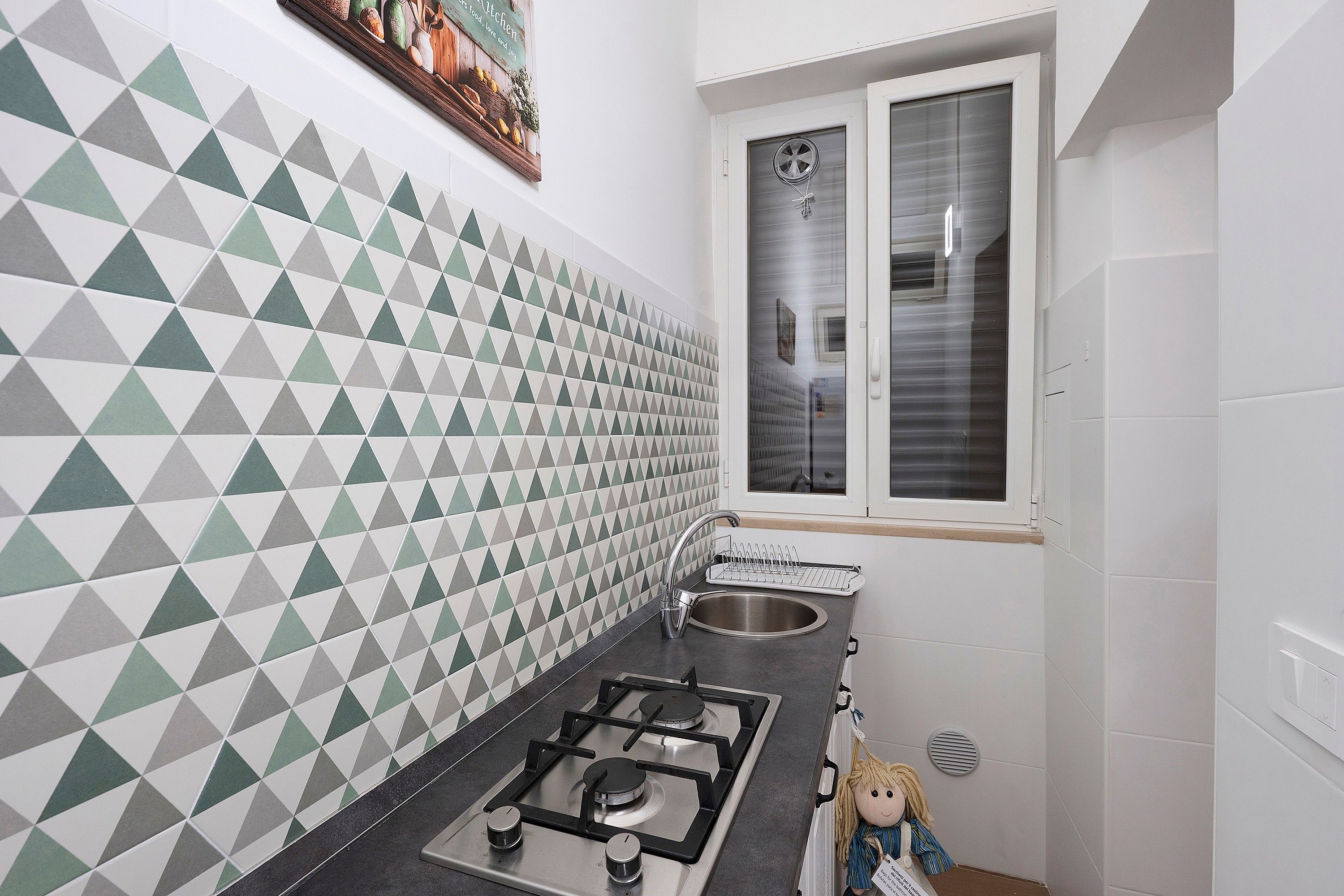 Een keuken met een achterwand met een geometrisch patroon, een raam met gordijn, een roestvrijstalen spoelbak en een gasfornuis.