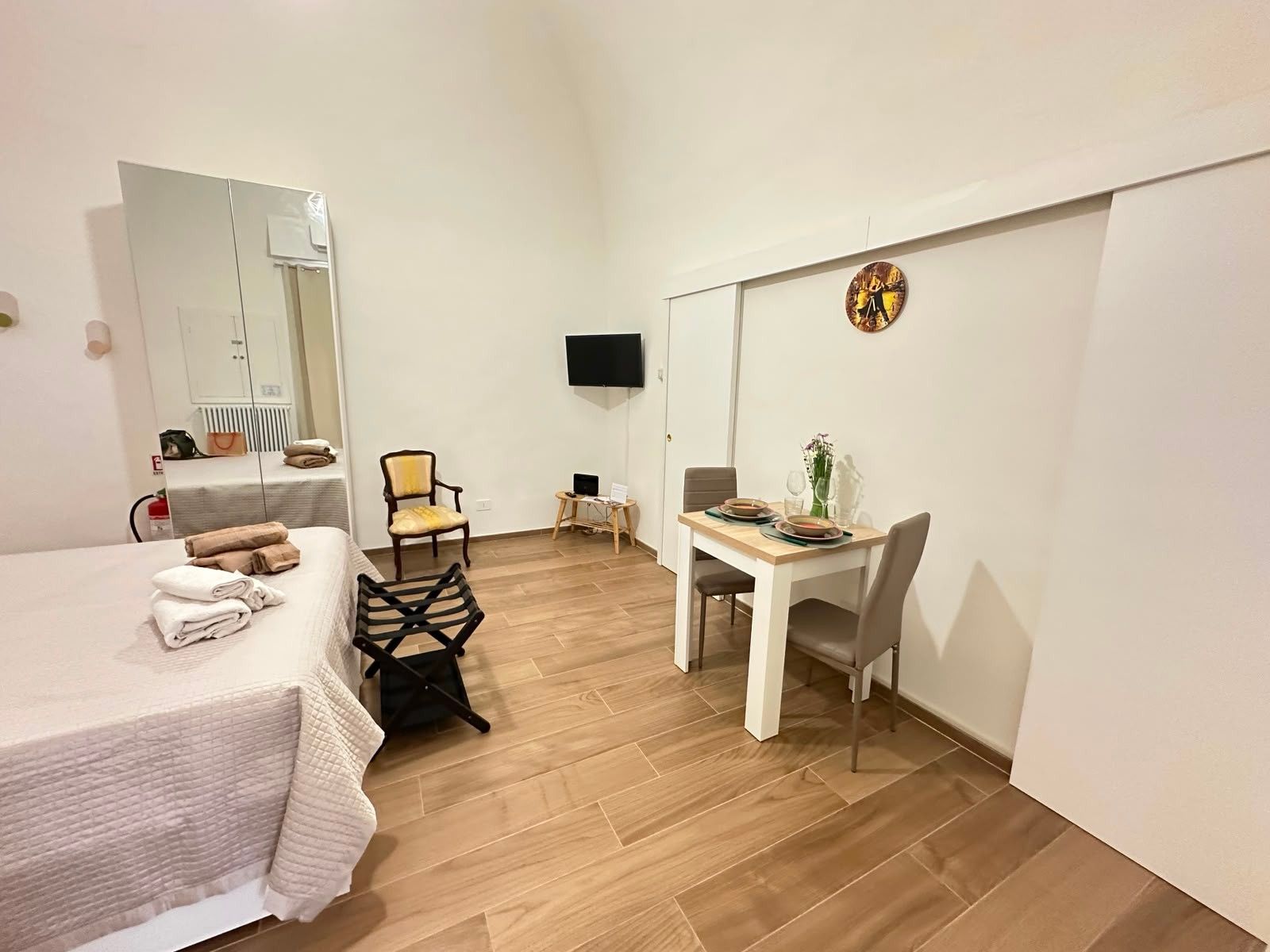 Una camera d'albergo con un letto, una sedia, un tavolino con un vaso, una TV a schermo piatto e una sveglia appesa al muro.