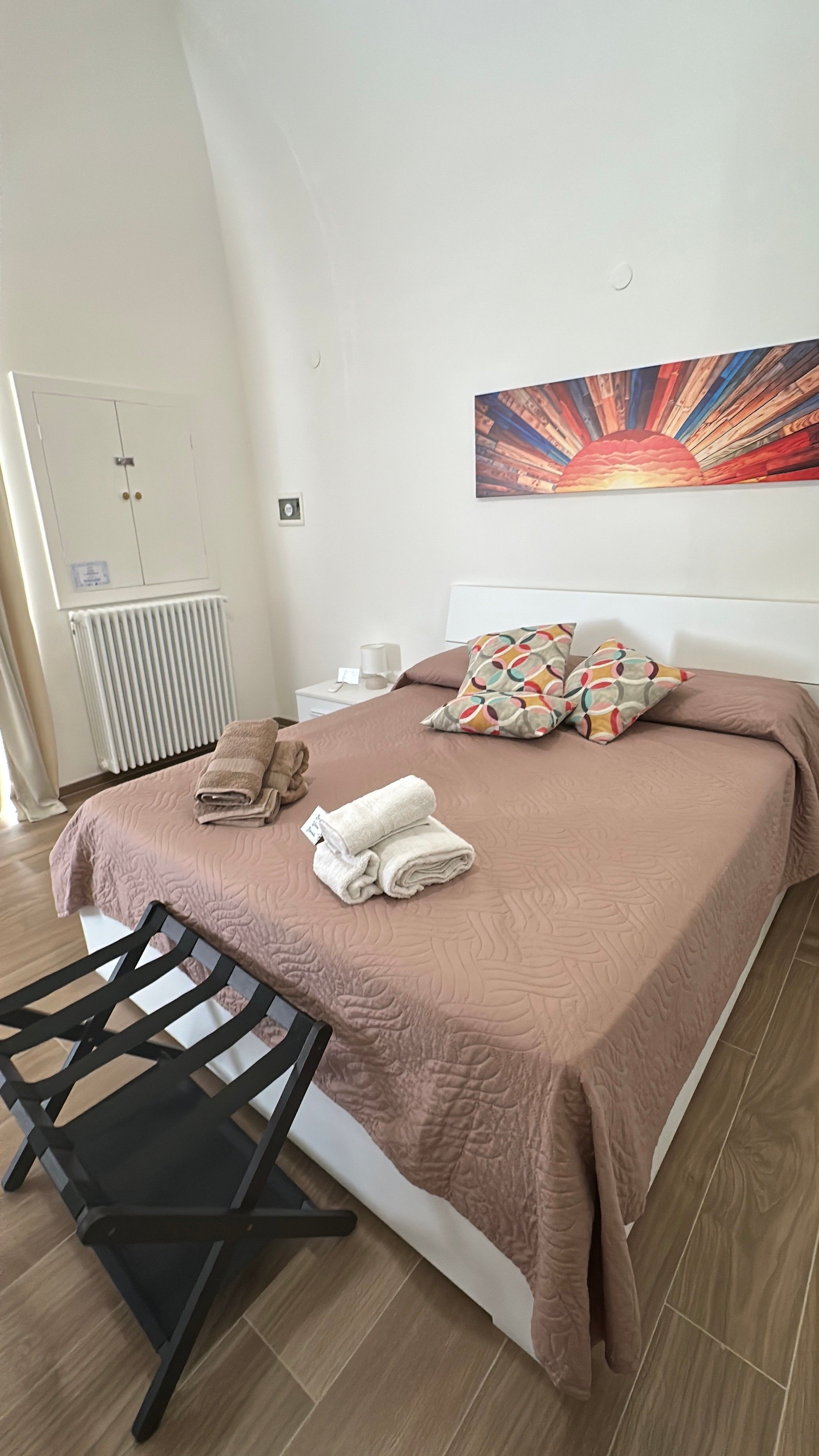 Un letto rifatto con cuscini e asciugamani piegati, un comodino con una lampada e un termosifone.