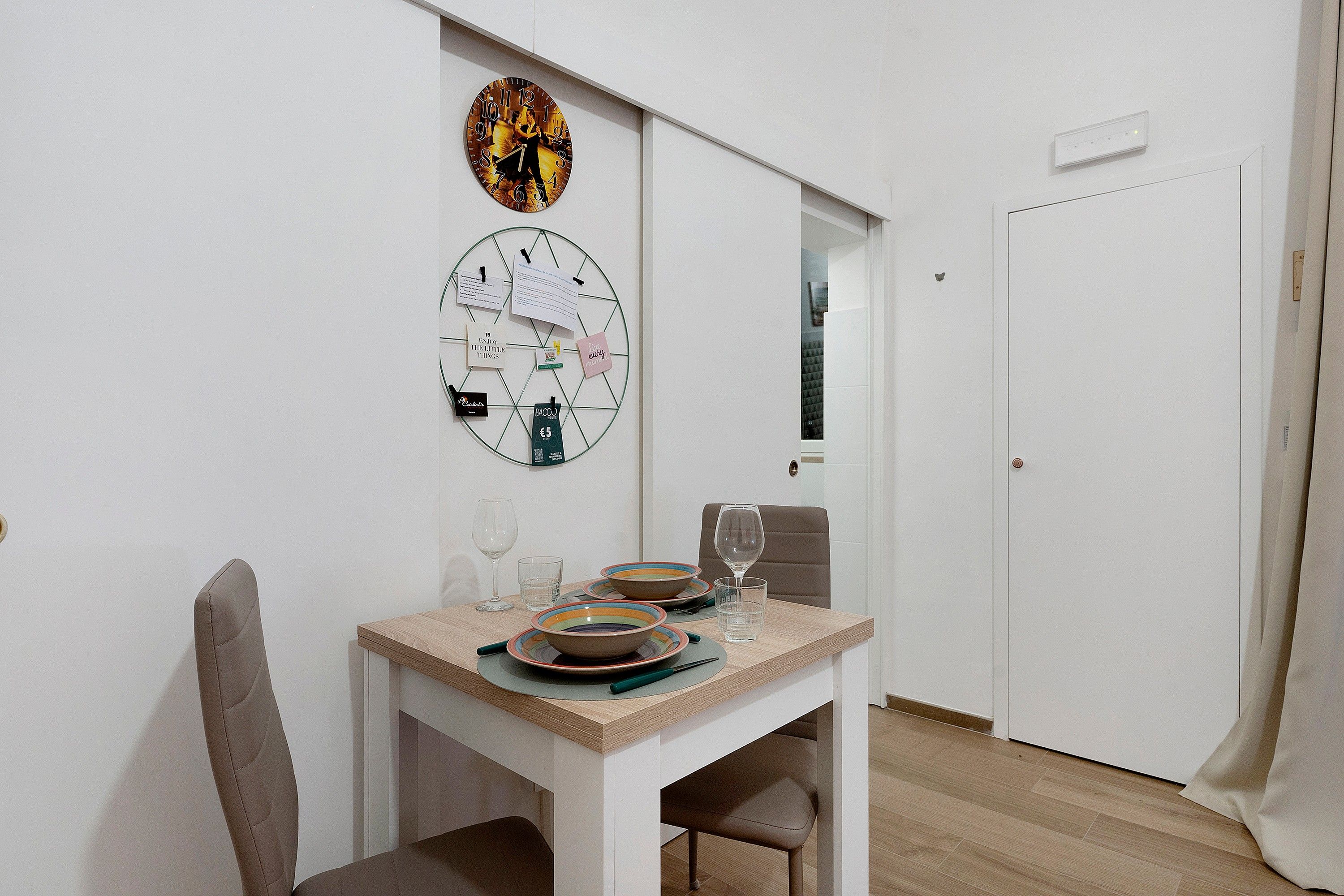 Een eetgedeelte met een tafel gedekt voor twee personen, een wandklok en een deur.