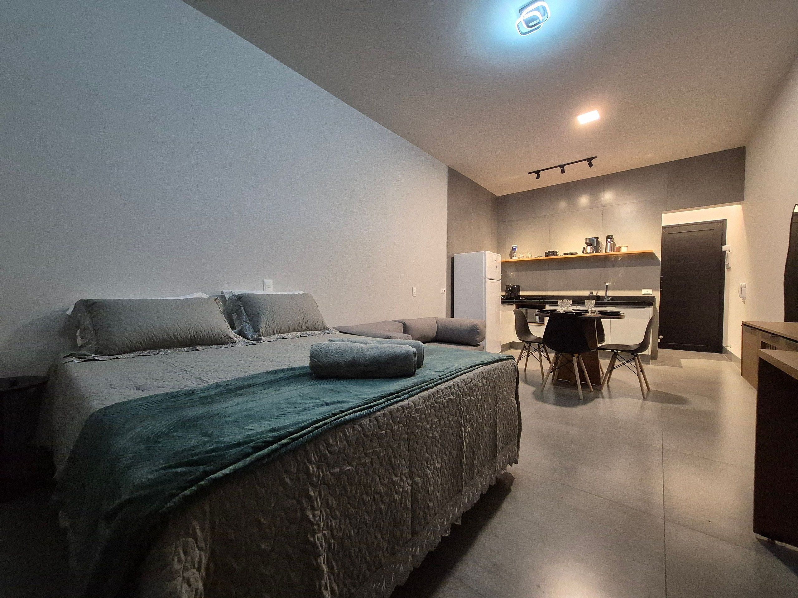 A modern bedroom with a bed, bedside tables, and a kitchenette in the background.