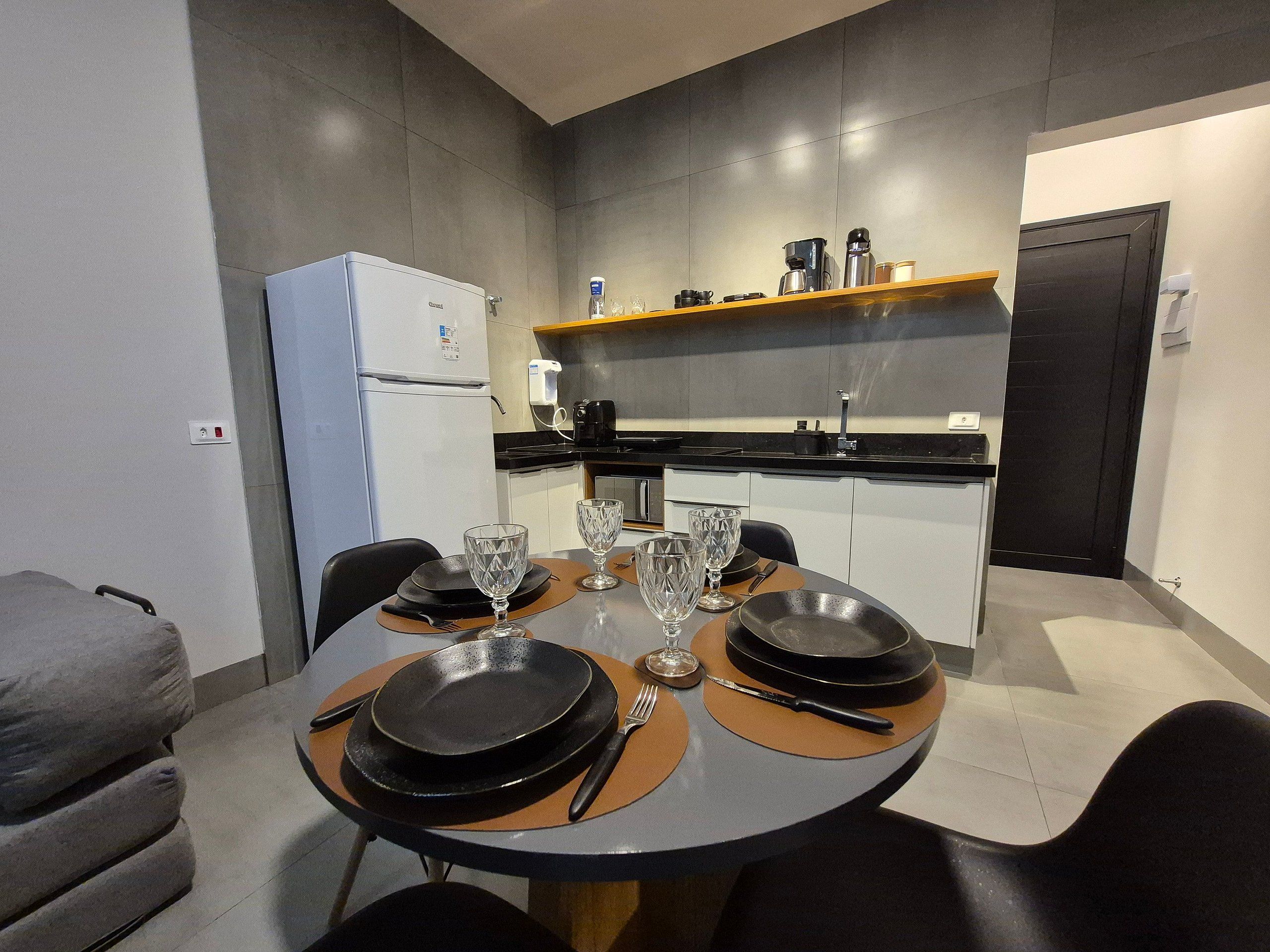 A modern kitchen with a dining area, equipped with a refrigerator, coffee maker, and a shelf with kitchen items.