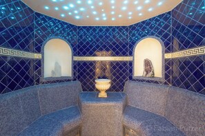 A room with a blue-tiled floor, two sofas, a central table, and a ceiling with circular lights.