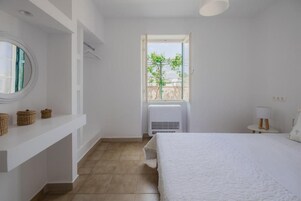 Un dormitorio con cama, ventana, radiador, espejo redondo y repisa con cestas.