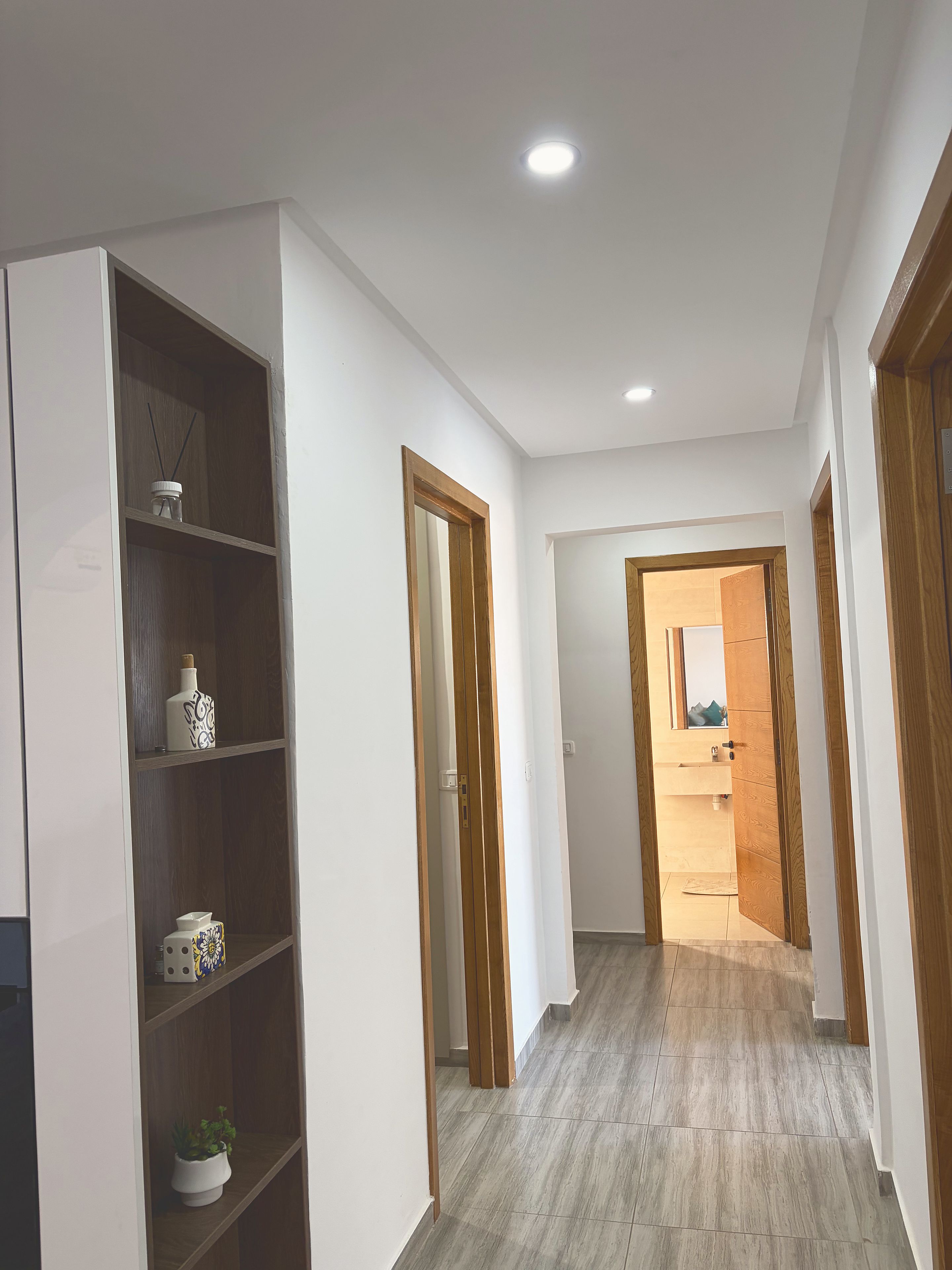 A hallway with a white wall, a wooden door frame, and a shelf with decorative items.
