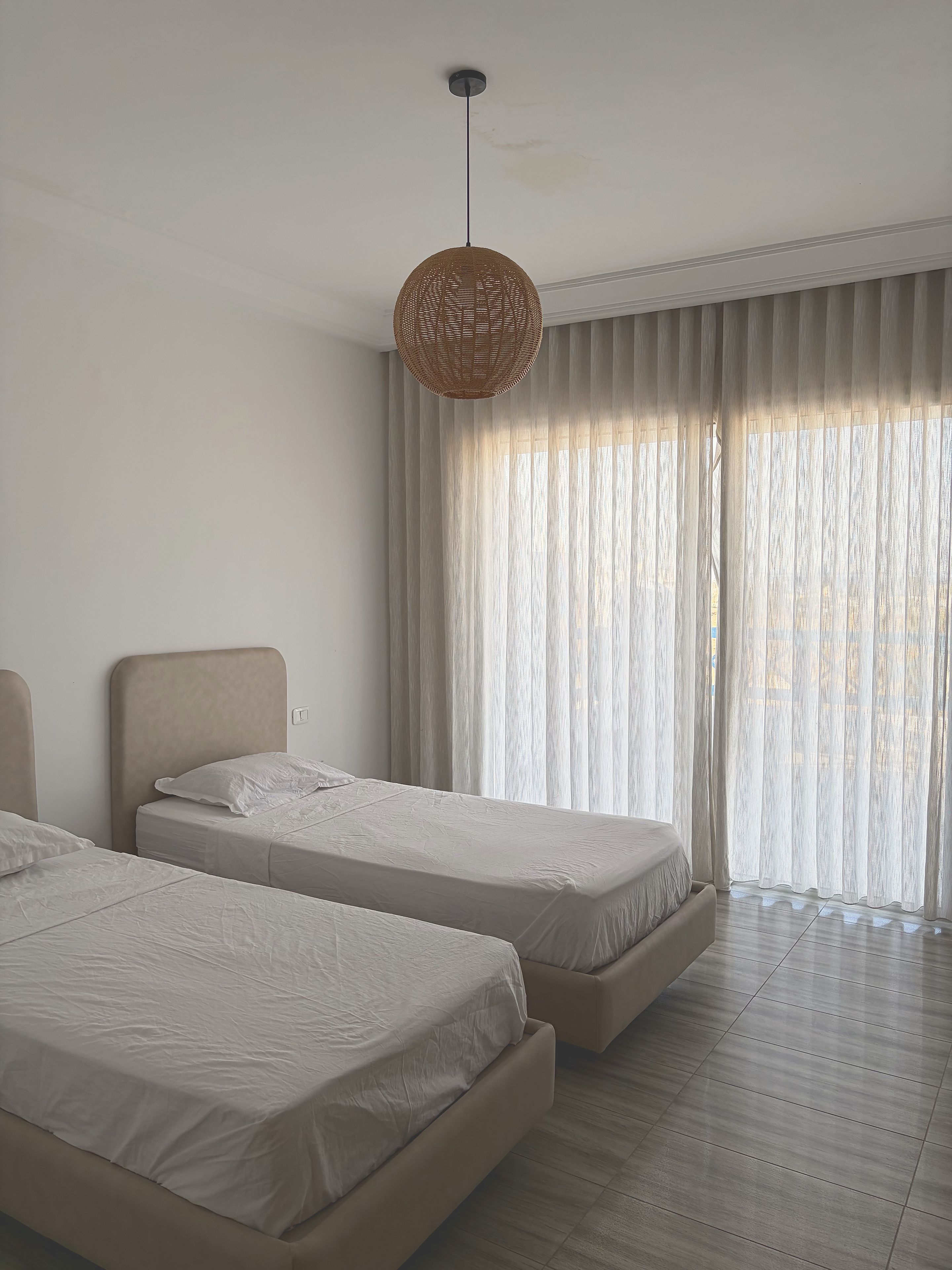 Un dormitorio con dos camas, una lámpara colgante y un ventanal con cortinas traslúcidas.