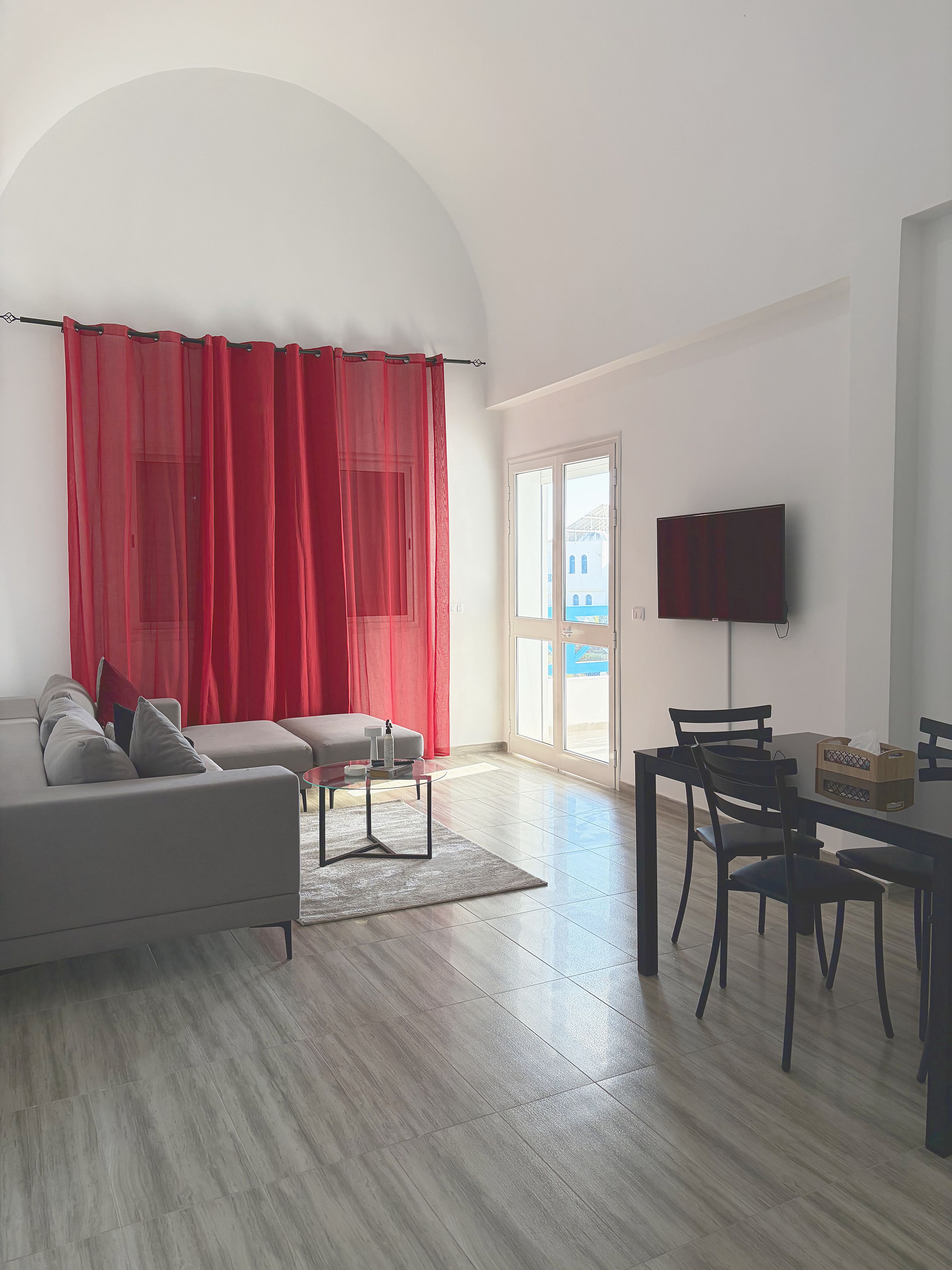A living room with a sofa, a coffee table, a dining table with chairs, a TV mounted on the wall, and a large window with a view.