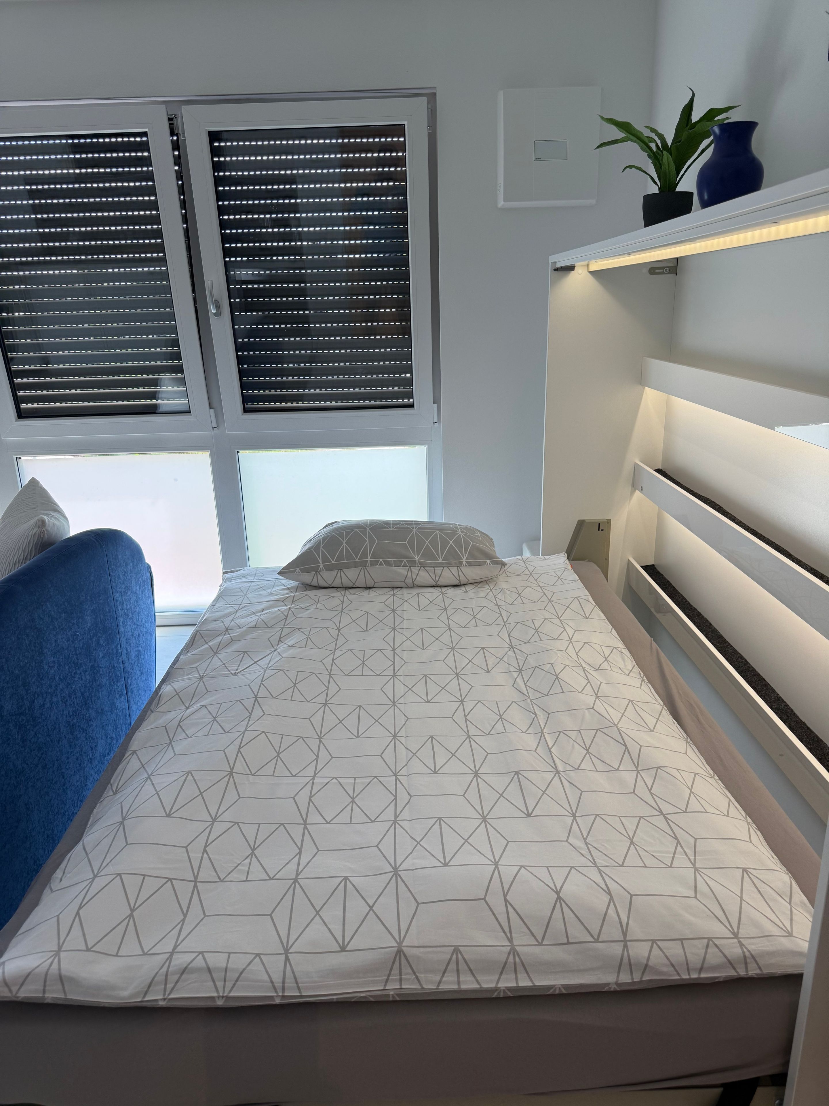 Une chambre à coucher avec un lit, une chaise bleue, une fenêtre avec des stores et une étagère sur laquelle se trouvent une plante et un vase.