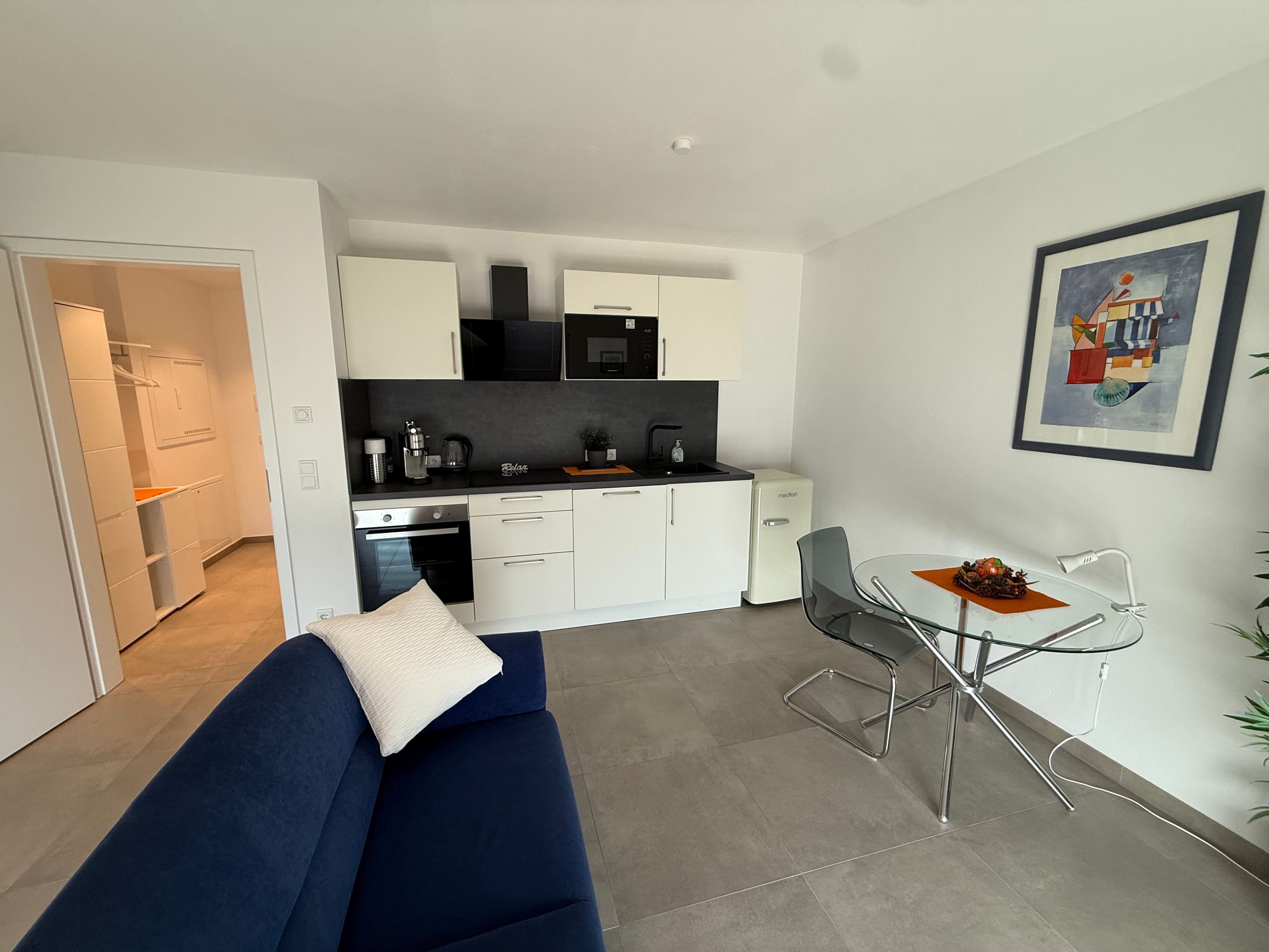 Un salon moderne avec un canapé bleu, une table à manger avec des chaises, un coin cuisine avec des meubles blancs et un tableau encadré au mur.