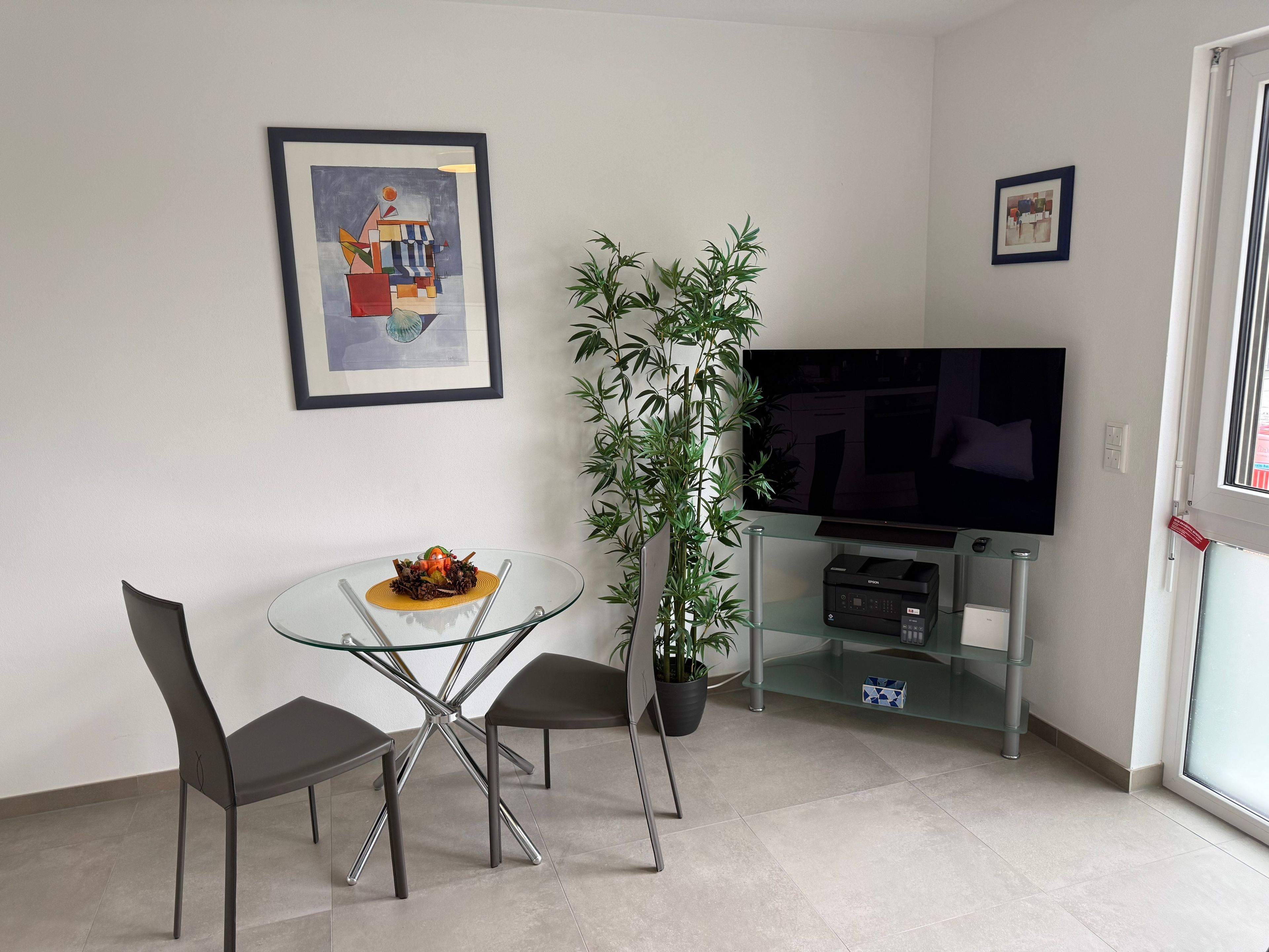A modern living room with a glass dining table, two chairs, a potted plant, a flat-screen TV, and artwork on the walls.