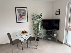 En moderne stue med et glasbord, to stole, en potteplante, et fladskærms-tv og kunst på væggene.