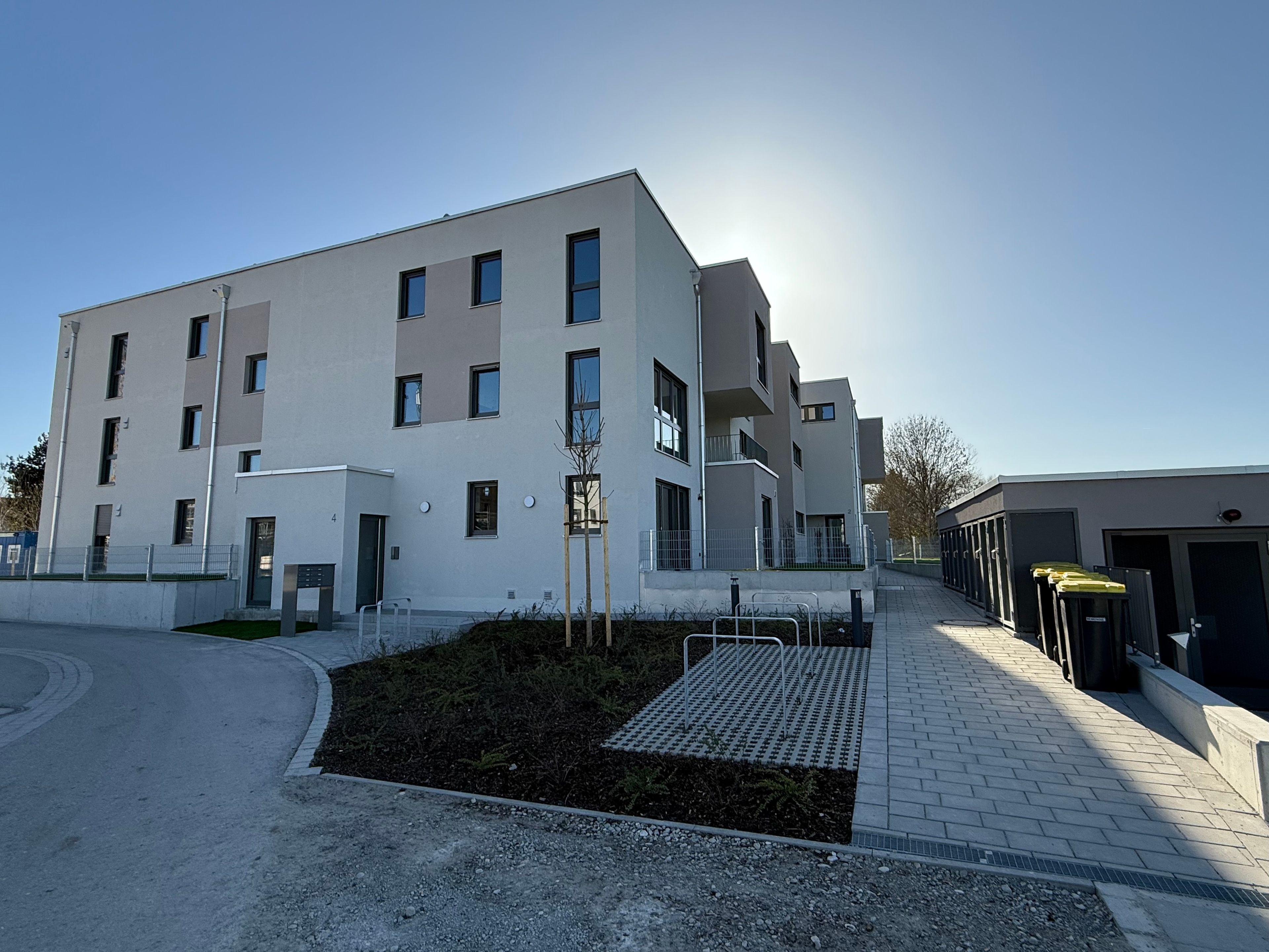 A modern building with multiple floors, a flat roof, and a paved walkway leading to a gated entrance.