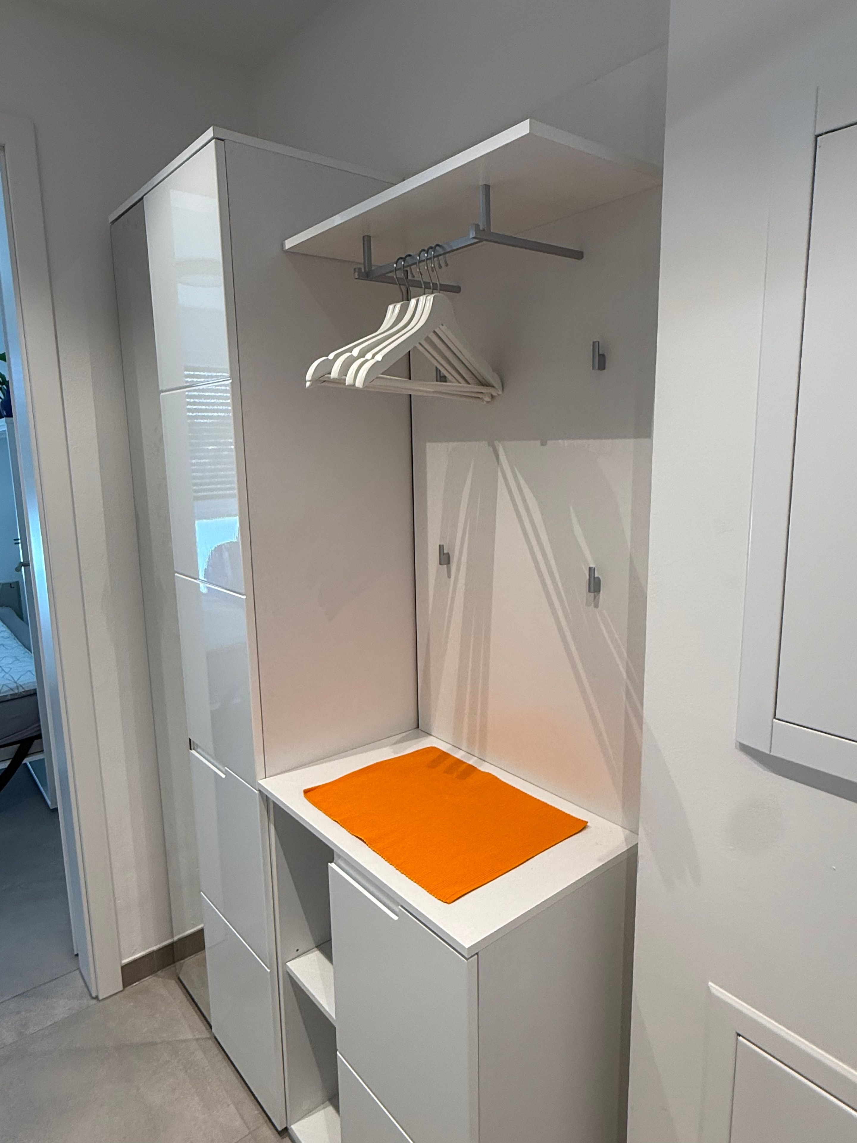 A compact room with a built-in wardrobe, a small seating area with an orange cushion, and a clothing rack.