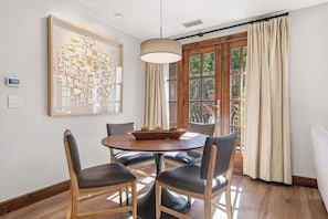 A dining area with a round table, four chairs, a wall-mounted light fixture, a window with curtains, and a framed abstract artwork. - Ascent 401 - 3 Bedroom Residence (Avon)