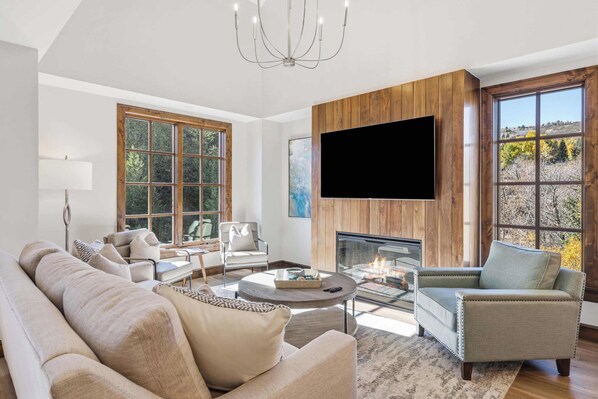 A spacious living room with a fireplace, large windows, and a flat-screen TV mounted on a wooden wall. - Ascent 401 - 3 Bedroom Residence (Avon)