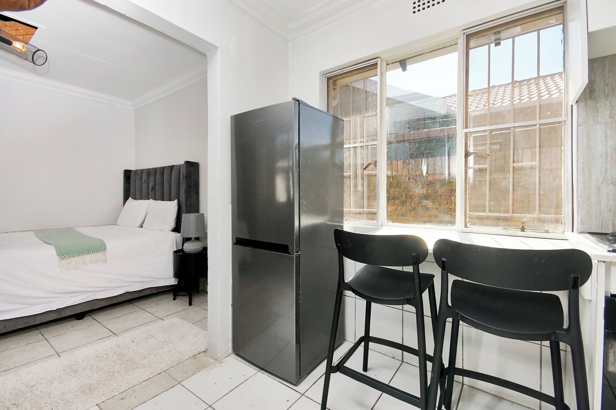 A compact room with a bed, a refrigerator, two chairs, and a window with a view of buildings.