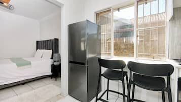 A compact room with a bed, a refrigerator, two chairs, and a window with a view of buildings.