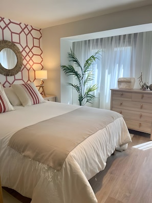 Un dormitorio con una cama grande, una mesita de noche con lámpara, un aparador y una ventana con cortinas traslúcidas.