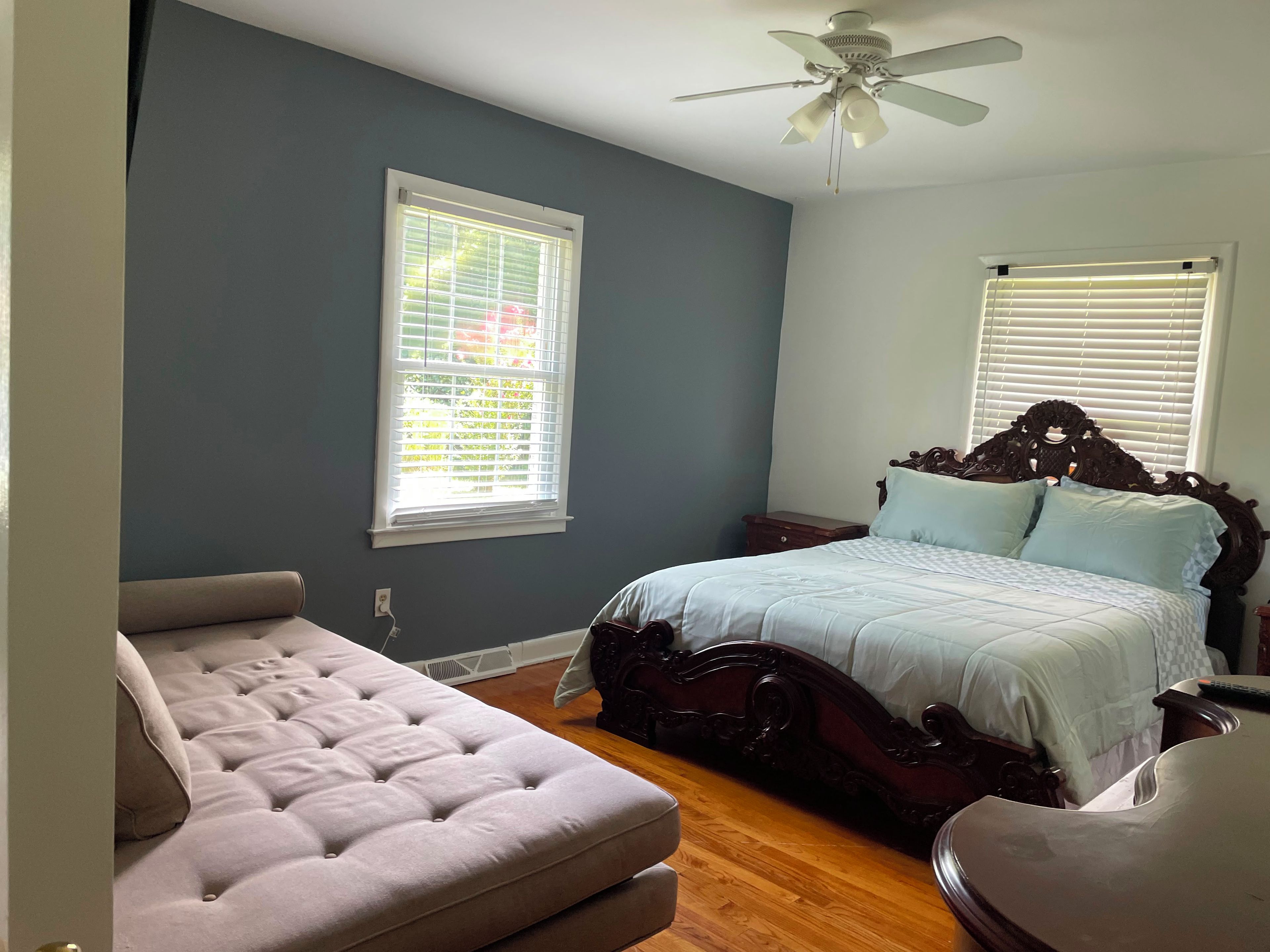A bedroom with a bed, a chair, a nightstand, a ceiling fan, and two windows with blinds.