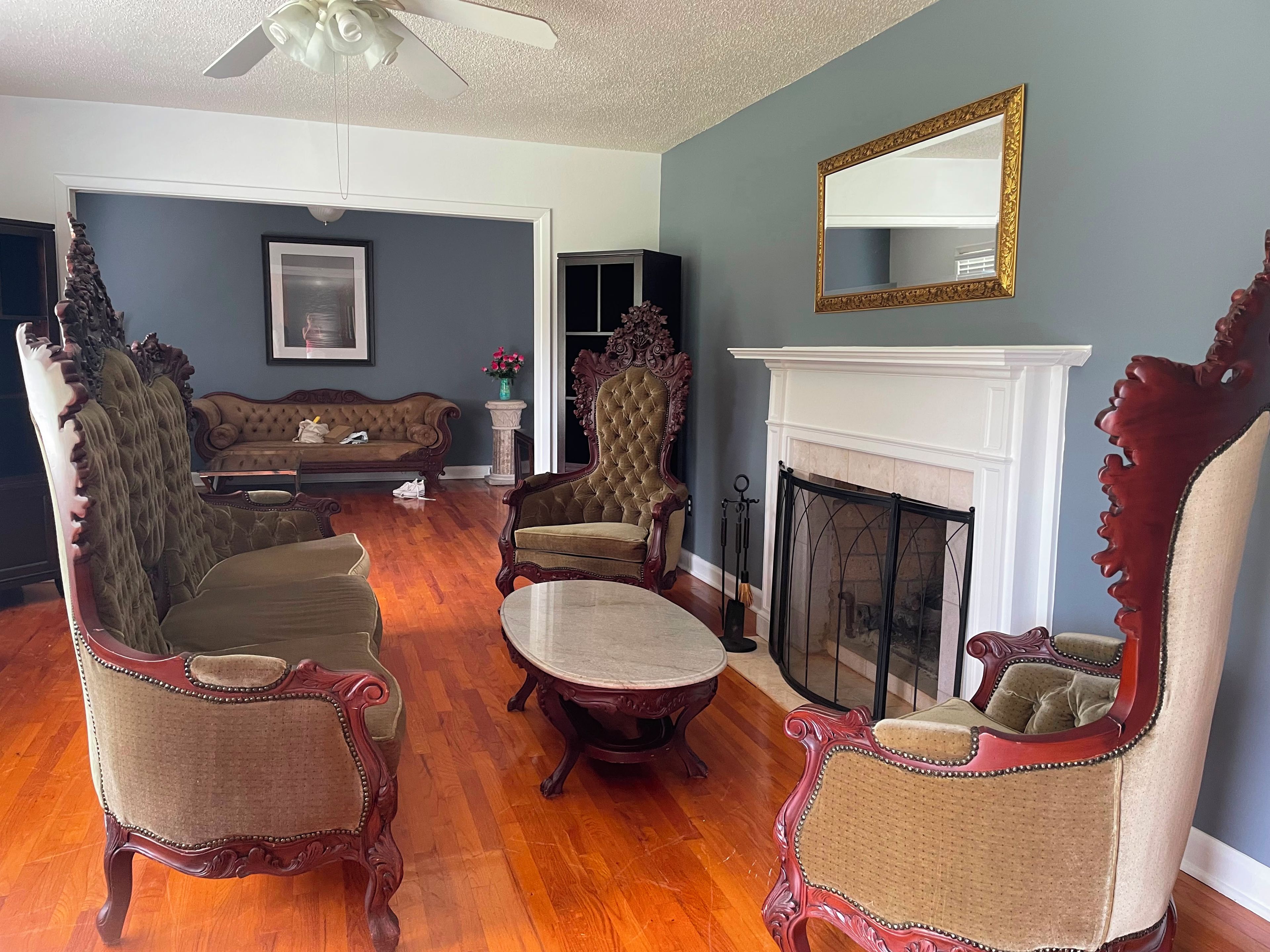 A living room with a fireplace, ornate chairs, a sofa, and a ceiling fan.