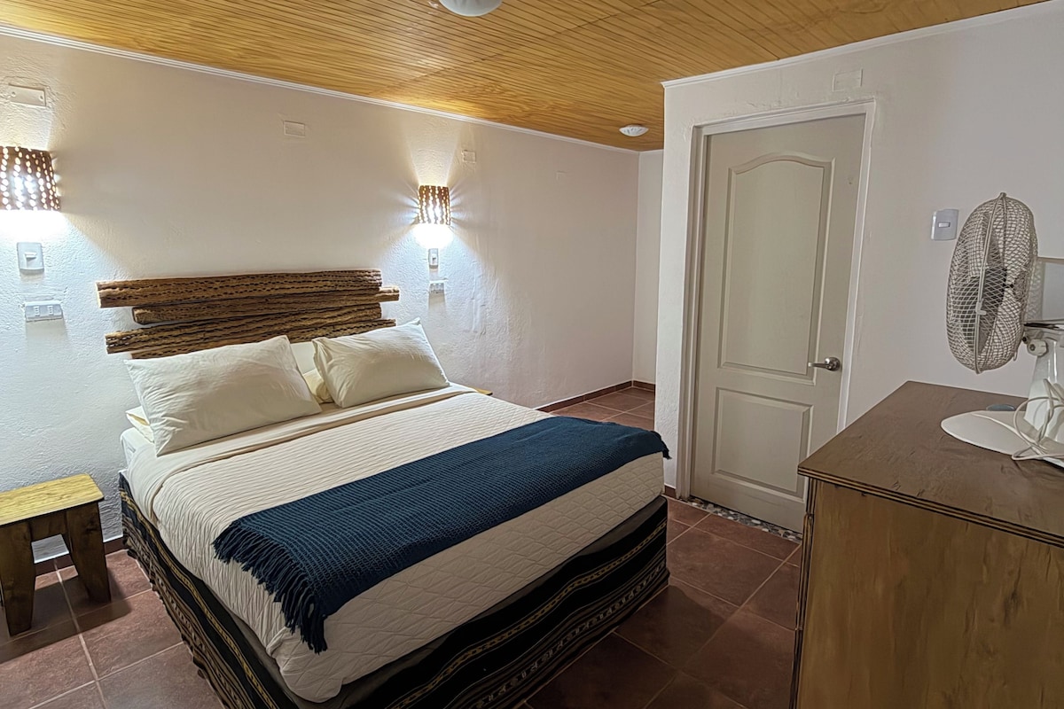 A bedroom with a wooden ceiling, a bed with a patterned bedspread, a bedside table, a door, and a small wooden cabinet.