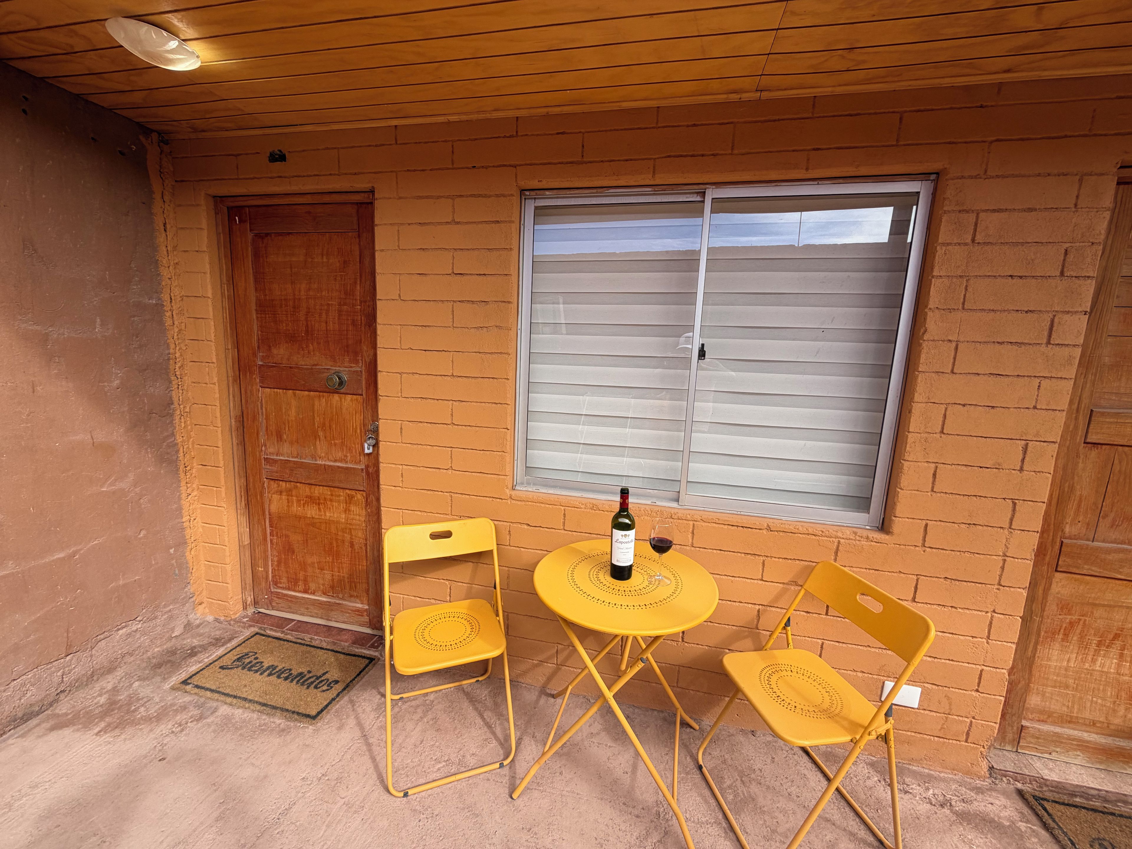 Una pequeña zona de estar al aire libre con una mesa, dos sillas, una botella de vino y una ventana con persianas.