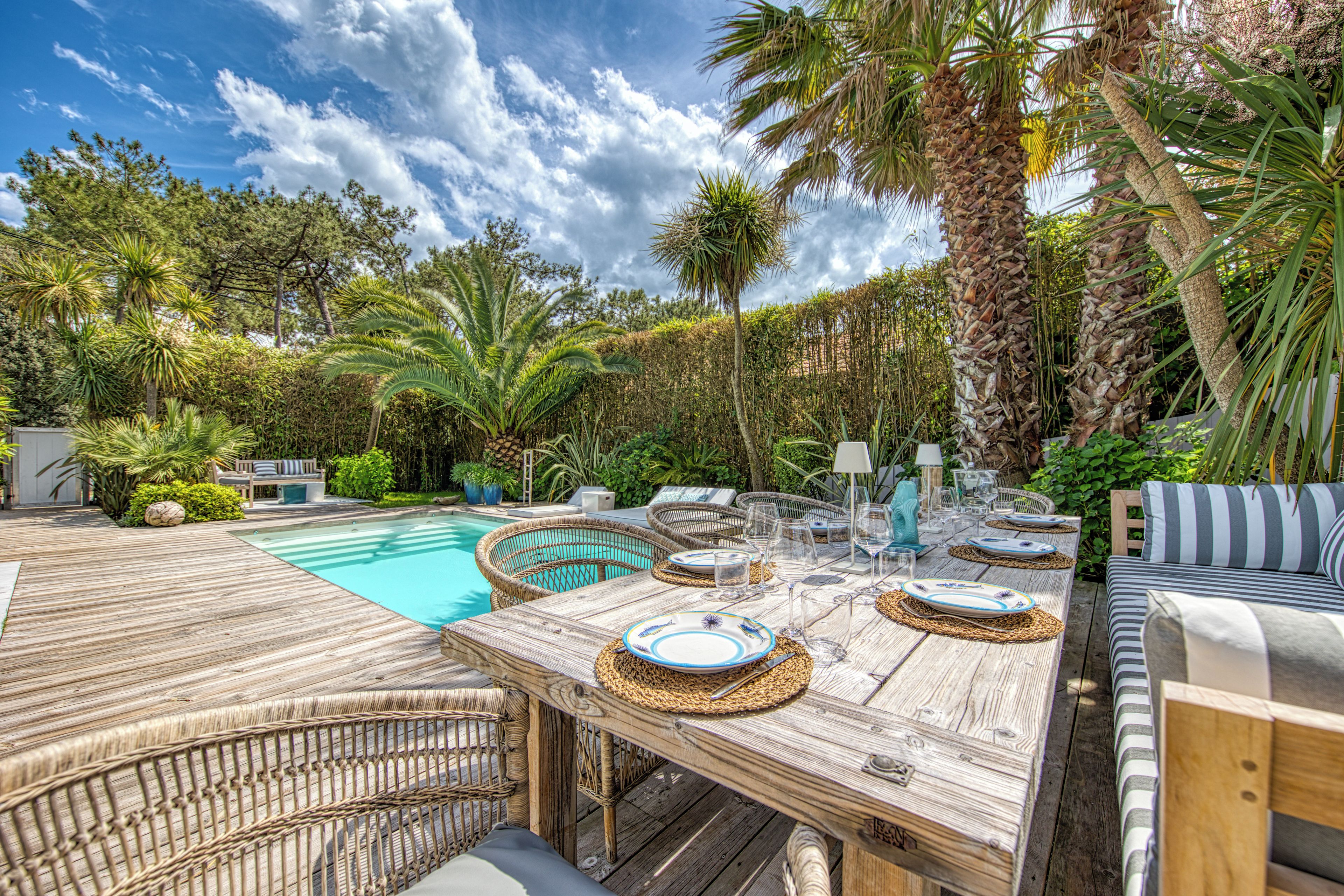 Een buitenzwembad met een houten terras, rieten meubels en een eettafel.