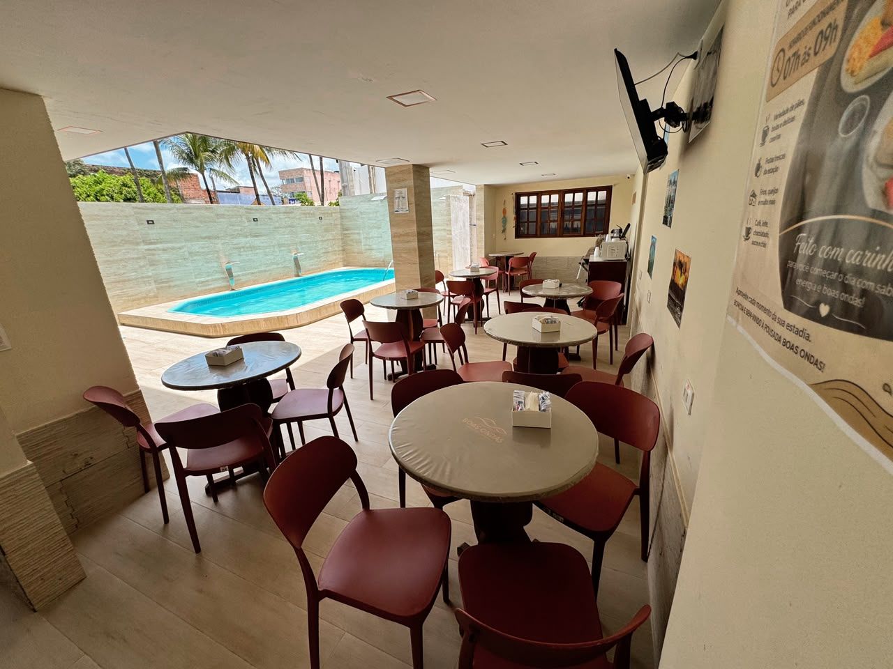 An indoor dining area with tables and chairs, a pool visible through a glass wall, and a poster on the wall.