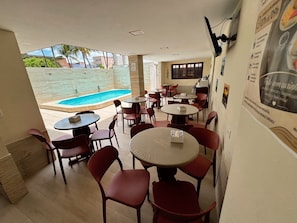 An indoor dining area with tables and chairs, a pool visible through a glass wall, and a poster on the wall. - Pousada Boas ondas (Ipojuca)