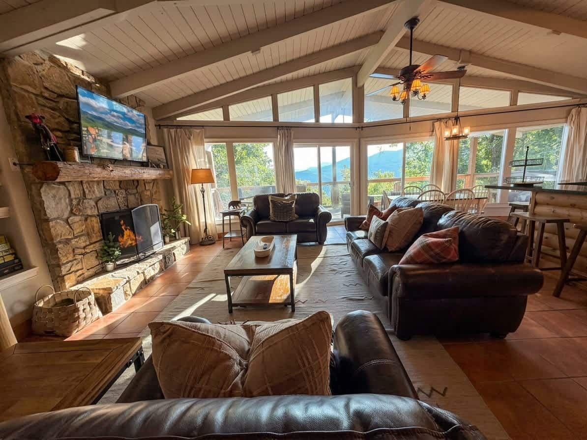Amplia sala de estar con chimenea de piedra, sofás de cuero, un televisor y ventanas grandes que ofrecen vistas al exterior.