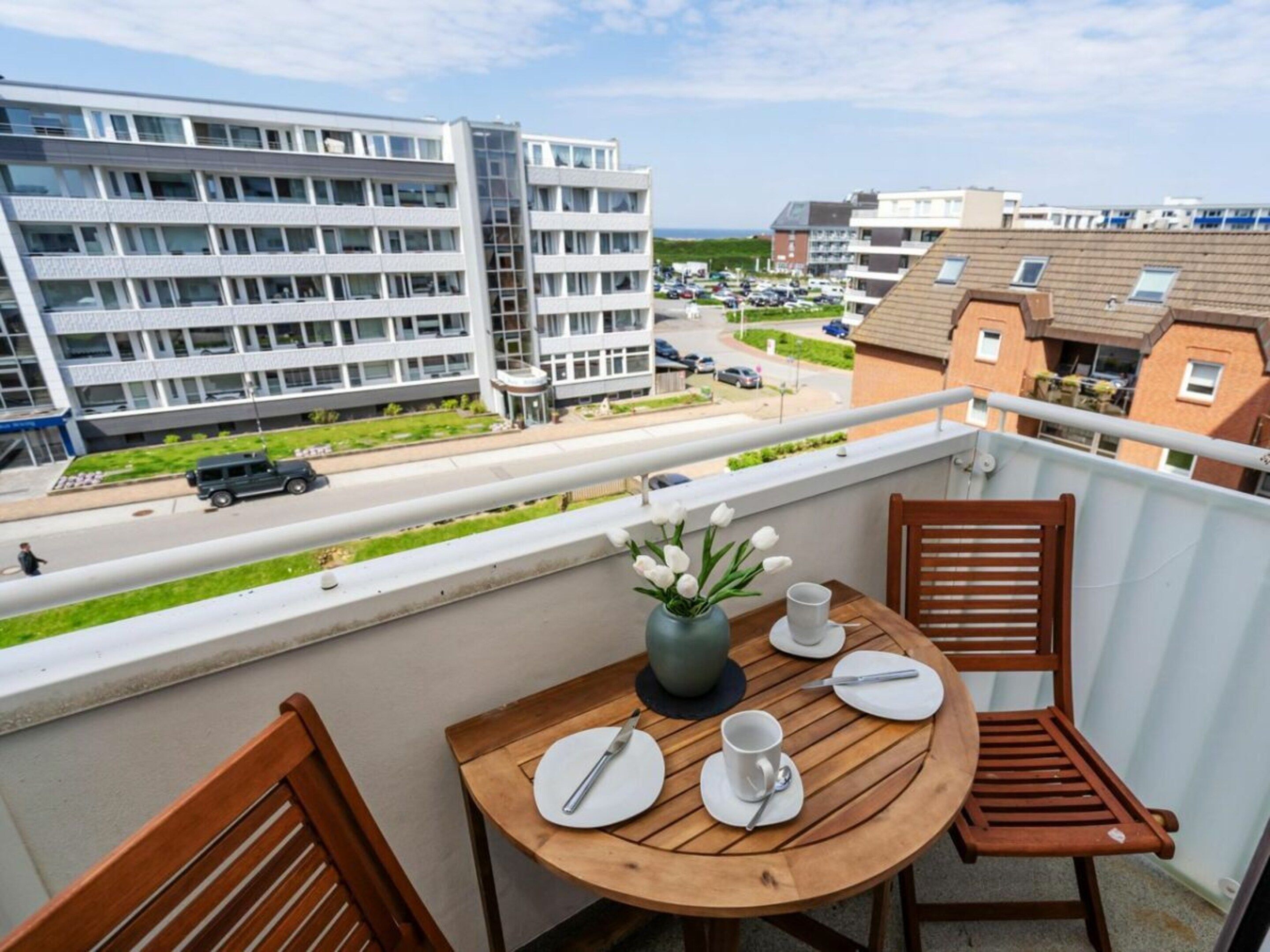 A balcony with a wooden table set for two, a vase with flowers, and a view of a multi-story building and a road with cars.
