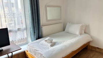 A single-bed room with a wooden frame, a white mattress, and a white pillow. There is a small table with a laptop, a window with blue curtains, and a wall-mounted mirror.