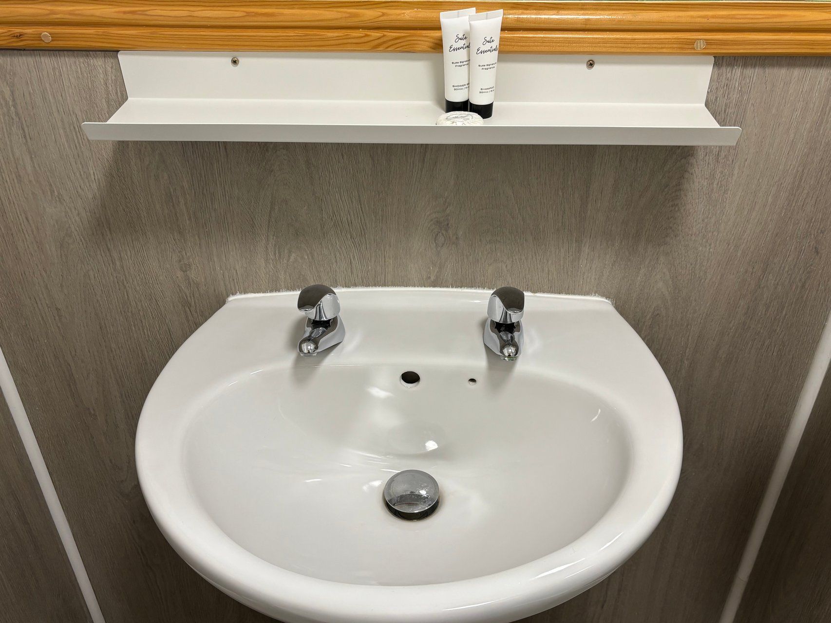 Un lavamanos blanco con dos grifos cromados, un estante de madera encima con artículos de tocador, y una pared de madera.
