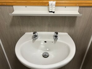 Un lavamanos blanco con dos grifos cromados, un estante de madera encima con artículos de tocador, y una pared de madera.