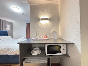 Habitación de hotel con cama, mesita de noche con microondas, tetera y bandeja con tazas y vaso.