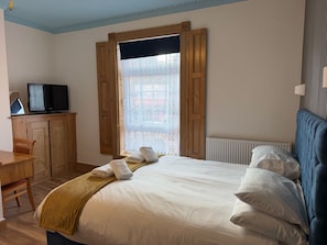 Een slaapkamer met een bed, een televisie op een houten kast, een raam met houten luiken en een radiator.
