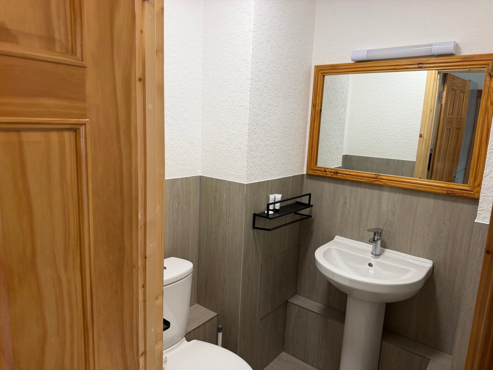 Een badkamer met een houten deur, een wit toilet, een wastafel en een spiegel met houten lijst.