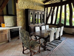 Un comedor con una mesa de madera, sillas blancas, una silla estampada, un armario y una zona de cocina con estufa y salpicadero de azulejos.