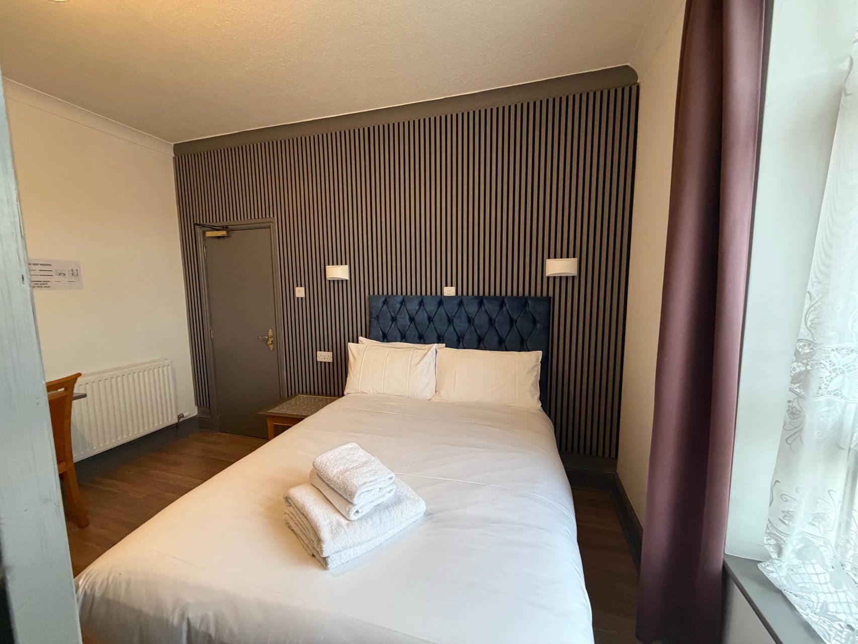 Habitación de hotel con una cama grande, cabecera, dos lámparas de noche, radiador y una ventana con cortinas.
