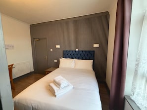 Et hotelværelse med en stor seng, sengegavl, to sengeborde med lamper, en radiator og et vindue med gardiner.