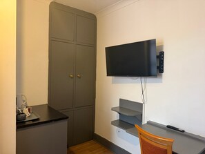 Et kompakt værelse med et fladskærms TV på væggen, en lille træstol og et tekøkken med en bordplade.