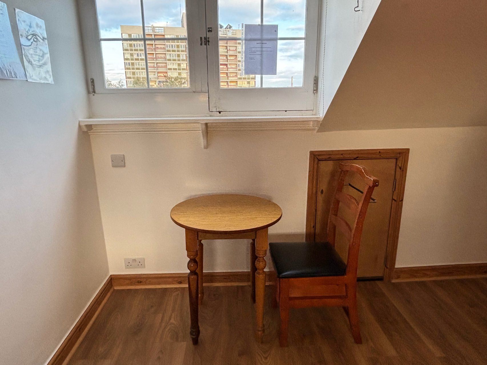 A room with a wooden table and chair, a window with a view of buildings, and a door.