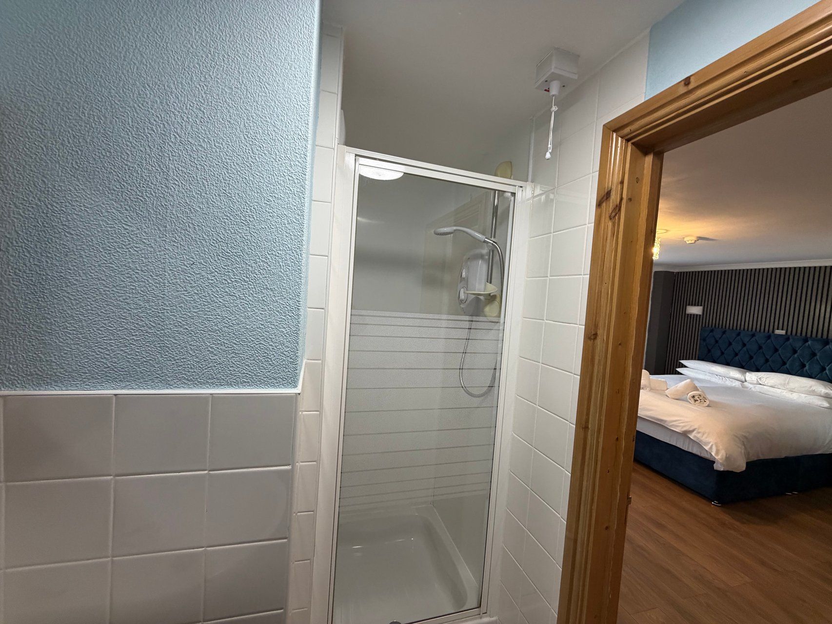 A hotel room with a glass-enclosed shower, a bed with a blue headboard, and a wooden floor.