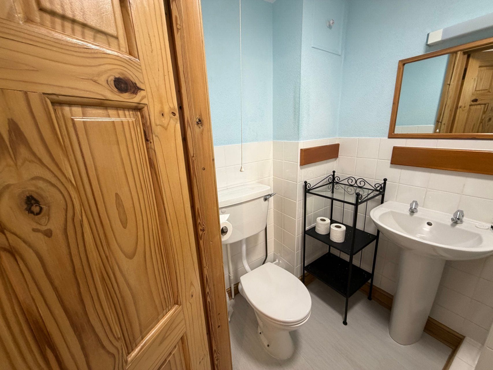 Un baño con puerta de madera, inodoro, lavabo y repisa con papel higiénico.